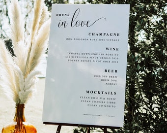 May include: A white sign with black text on an easel, listing drinks for a wedding reception. The sign reads "Drunk in love" and lists champagne, wine, beer, and mocktails. The sign is in front of a brown bottle with a label that says "Cabernet".