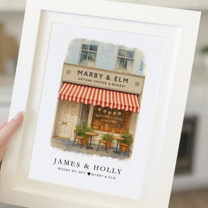 May include: A framed print featuring a watercolor illustration of a coffee shop with a red and white striped awning. The shop is named "Marby & Elm, Artisan Coffee & Bakery." The print also includes the text "James & Holly, Where We Met Marby & Elm."
