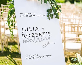 以下が含まれることがあります： 白い結婚式の看板に、黒い文字で「Welcome to the celebration of Julia + Robert's wedding」と「22.06.2022 Cotton Beach Club」と書かれています。看板は白いイーゼルに置かれ、緑の葉で飾られています。