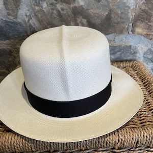 Pu&ograve; includere: Un cappello di paglia bianco con un nastro nero. Il cappello &egrave; appoggiato su un cesto intrecciato.