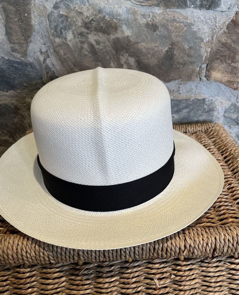 Pu&ograve; includere: Un cappello di paglia bianco con un nastro nero. Il cappello &egrave; appoggiato su un cesto intrecciato.