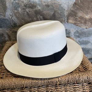 Pu&ograve; includere: Un cappello Panama bianco con un nastro nero. Il cappello &egrave; appoggiato su un cesto intrecciato.