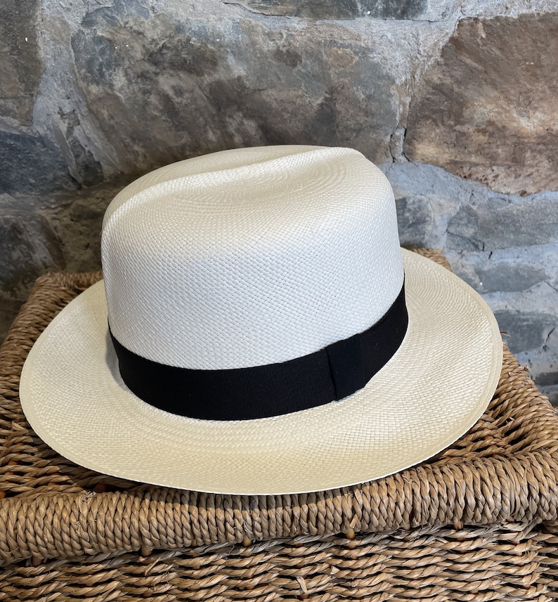 Pu&ograve; includere: Un cappello Panama bianco con un nastro nero. Il cappello &egrave; appoggiato su un cesto intrecciato.
