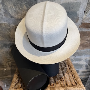 Pu&ograve; includere: Un cappello Panama bianco con un nastro nero. Il cappello &egrave; appoggiato su un cesto di vimini.