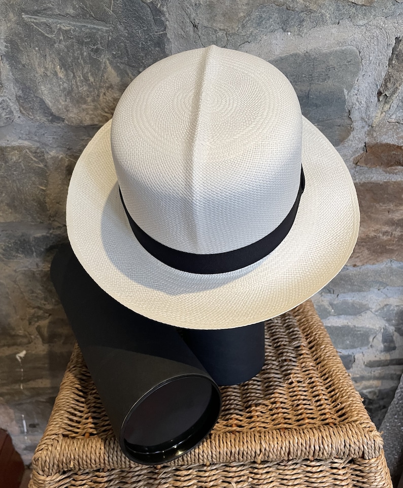 Pu&ograve; includere: Un cappello Panama bianco con un nastro nero. Il cappello &egrave; appoggiato su un cesto di vimini.