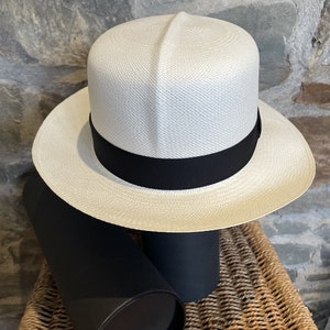 Pu&ograve; includere: Un cappello Panama bianco con un nastro nero. Il cappello &egrave; appoggiato su una scatola cilindrica nera.
