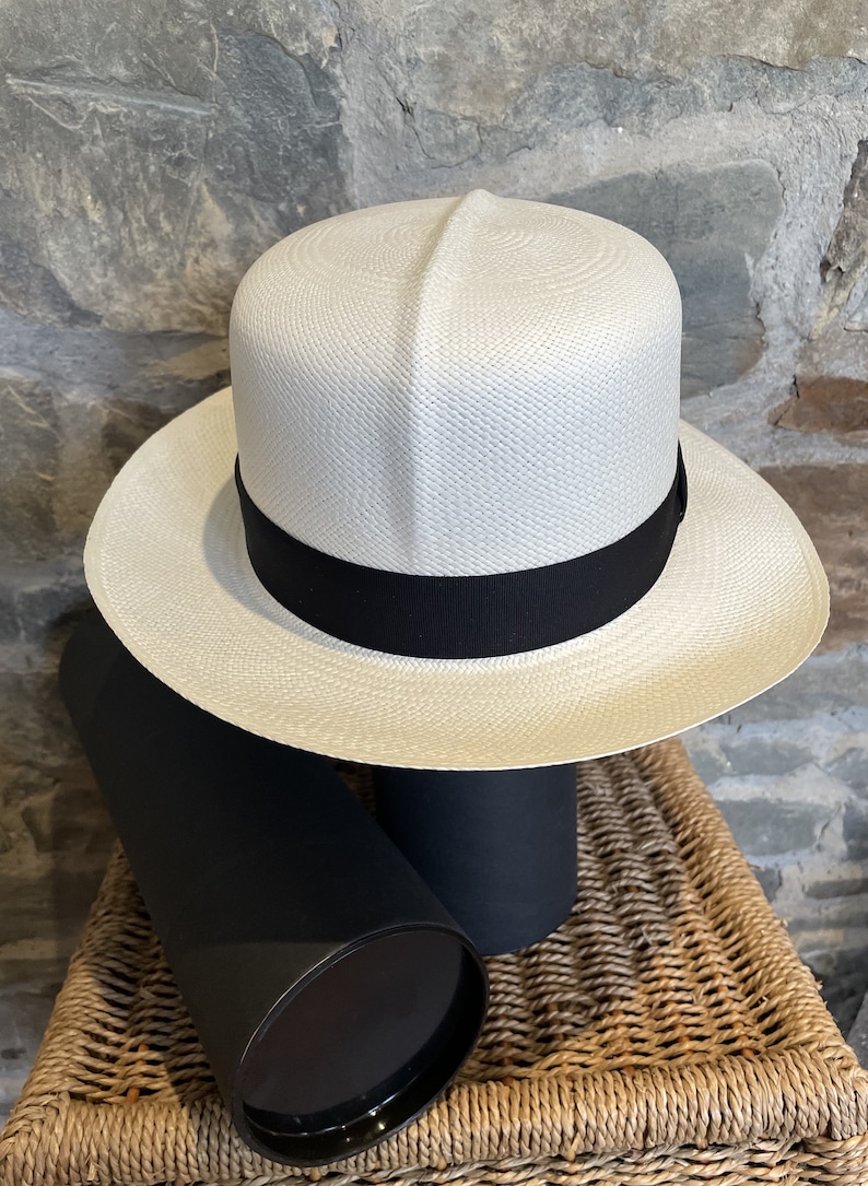Pu&ograve; includere: Un cappello Panama bianco con un nastro nero. Il cappello &egrave; appoggiato su una scatola cilindrica nera.