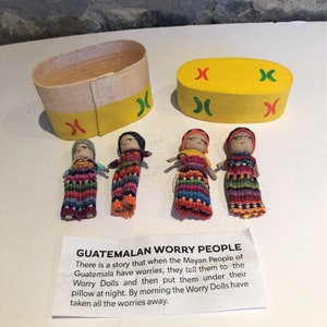 May include: Four small colourful worry dolls made of fabric and yarn, sitting on a white surface next to a yellow wooden box with red and green X's painted on it. The dolls are traditionally used in Guatemala to help people sleep by taking away their worries. A small card with the text "GUATEMALAN WORRY PEOPLE" and a description of the dolls' purpose is also visible.
