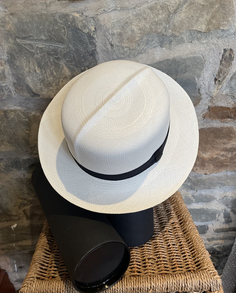 Pu&ograve; includere: Un cappello Panama bianco con un nastro nero. Il cappello &egrave; appoggiato su una scatola cilindrica nera e marrone.