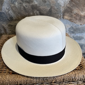Pu&ograve; includere: Un cappello Panama bianco con un nastro nero. Il cappello &egrave; appoggiato su un cesto intrecciato.