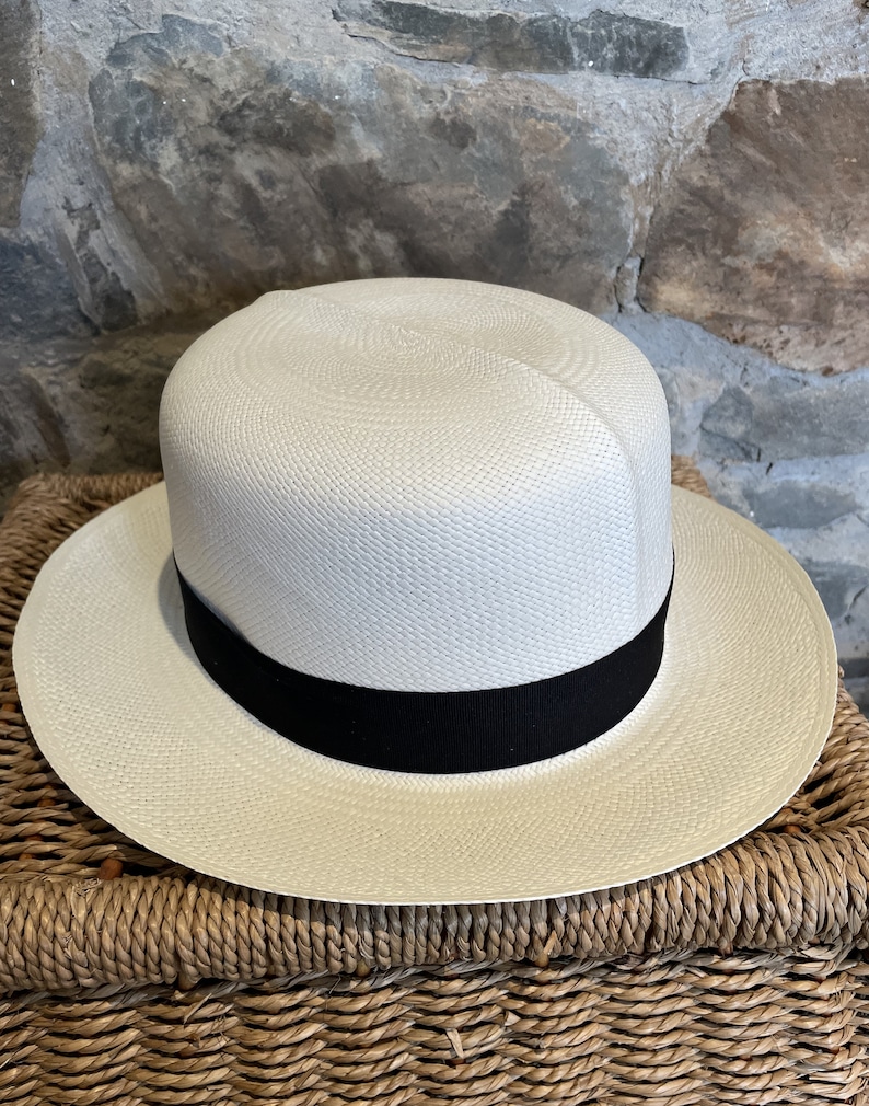 Pu&ograve; includere: Un cappello Panama bianco con un nastro nero. Il cappello &egrave; appoggiato su un cesto intrecciato.