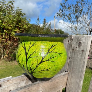 May include: A green and black ceramic planter with a tree branch design. The planter is sitting on a wooden fence.