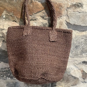 May include: A brown woven tote bag with thick, braided handles. The bag is made of a natural fibre and has a slightly textured surface.