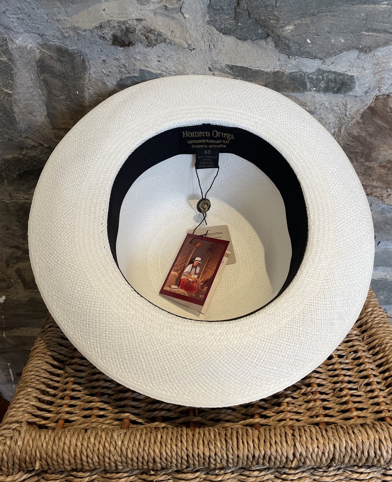 Pu&ograve; includere: Cappello Panama bianco con una fascia nera e un'etichetta che recita "Homero Ortega Genuine Panama Hat Cuenca-Ecuador" e "60".