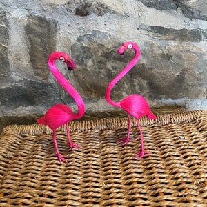 May include: Two pink flamingo figurines with long, curved necks and black eyes. The figurines are standing on a woven wicker surface.