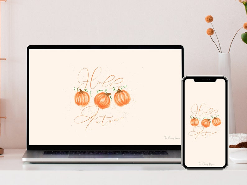 May include: A digital desktop and phone wallpaper featuring a light beige background with the text "Hello Autumn" in a cursive font. Three orange pumpkins with green stems are arranged in a row above the text. The text "The Cozy Hype" is in the bottom right corner of the image.