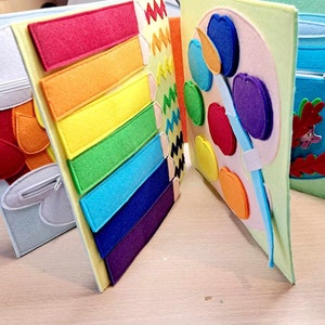 May include: A felt book with four pages. The first page has a red and orange felt mitten with a zipper. The second page has a rainbow of felt strips. The third page has a felt paint palette with colorful felt apples. The fourth page has a blue felt fishbowl with a zipper and a felt fish.