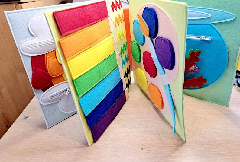 May include: A felt book with four pages. The first page has a red and orange felt mitten with a zipper. The second page has a rainbow of felt strips. The third page has a felt paint palette with colorful felt apples. The fourth page has a blue felt fishbowl with a zipper and a felt fish.