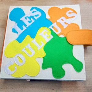 May include: A felt book for children with the words "Les Couleurs" in white on a yellow, orange, blue, and green background.