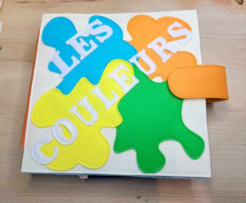 May include: A felt book for children with the words "Les Couleurs" in white on a yellow, orange, blue, and green background.
