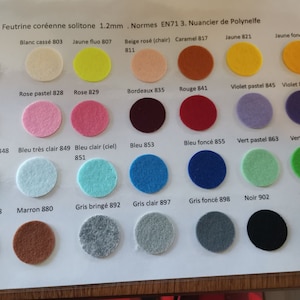 Puede incluir: Una carta de colores de muestras de tela de fieltro en varios colores, incluyendo blanco, beige, naranja, rosa, rojo, morado, azul, verde, marrón, gris y negro. Cada muestra está etiquetada con un nombre de color y un número.