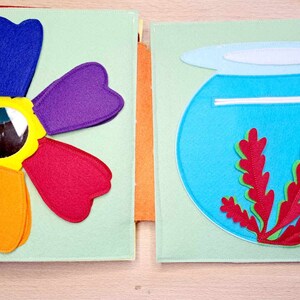 May include: A felt book with two pages. The left page features a rainbow flower with a mirror in the center. The right page features a blue felt fishbowl with a red felt seaweed and a pink felt fish.