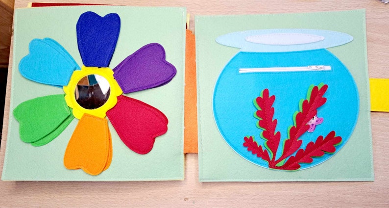 May include: A felt book with two pages. The left page features a rainbow flower with a mirror in the center. The right page features a blue felt fishbowl with a red felt seaweed and a pink felt fish.