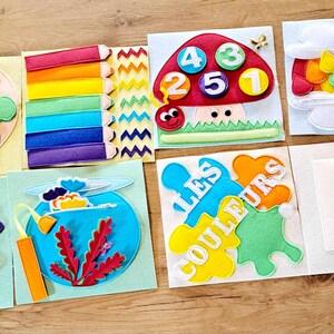 May include: A felt activity book for children with colorful pages featuring a paint palette, rainbow, flower, fishbowl, mushroom with numbers, rainbow in a cloud, and a red paint splatter. The book has the text "Les Couleurs" on one page.