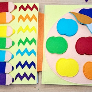 May include: A felt book with two pages. The left page has rainbow colored pencils with a zig-zag pattern in each color. The right page has a felt paint palette with six different colored paint pots and a blue paintbrush.