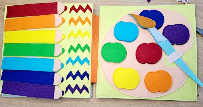 May include: A felt book with two pages. The left page has rainbow colored pencils with a zig-zag pattern in each color. The right page has a felt paint palette with six different colored paint pots and a blue paintbrush.