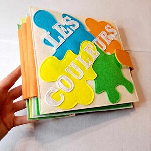 May include: A felt book for children with the title "Les Couleurs" in white letters on a yellow, green, blue and orange background.