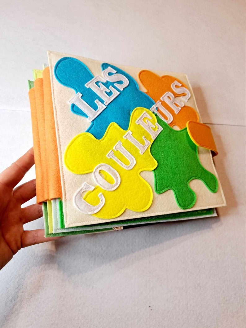May include: A felt book for children with the title "Les Couleurs" in white letters on a yellow, green, blue and orange background.