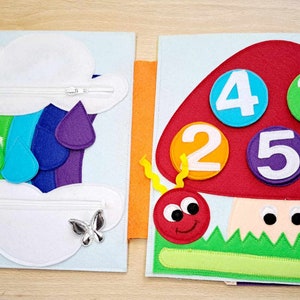 May include: A felt book with two pages. The left page features a rainbow with raindrops and a white cloud with a zipper. The right page features a red mushroom with a ladybug and numbers 1, 2, 3, 4, and 5.