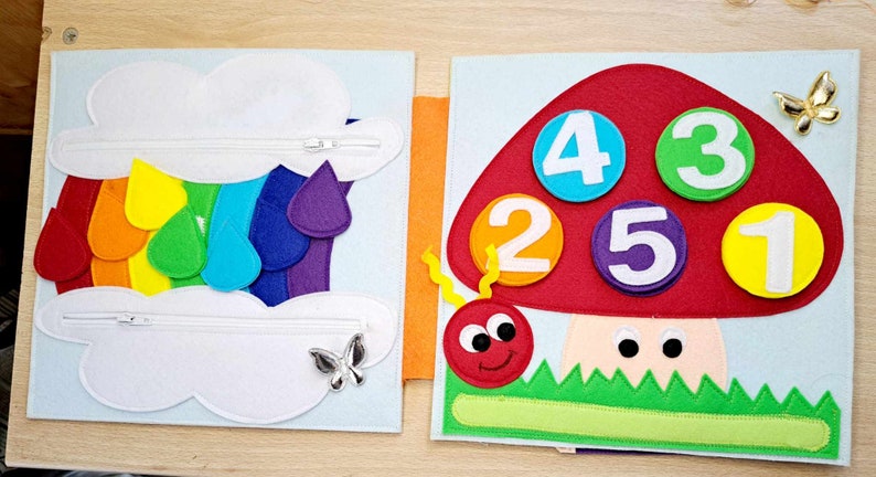 May include: A felt book with two pages. The left page features a rainbow with raindrops and a white cloud with a zipper. The right page features a red mushroom with a ladybug and numbers 1, 2, 3, 4, and 5.