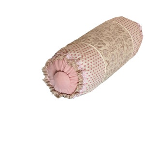 Puede incluir: Una almohada cilíndrica con estampado floral rosa y beige. La almohada tiene un borde de encaje y un botón rosa en el centro.