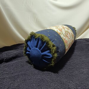 May include: A blue and white patterned cylindrical pillow with a green fringe trim. The pillow is on a dark blue surface.