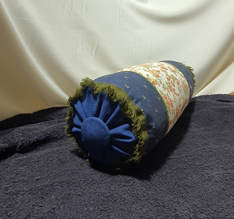 May include: A blue and white patterned cylindrical pillow with a green fringe trim. The pillow is on a dark blue surface.
