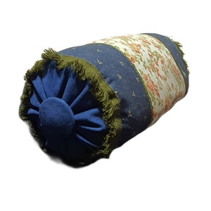 May include: A blue and white patterned cylindrical pillow with a green fringe trim. The pillow has a floral design on one side and a blue fabric with white dots on the other side.