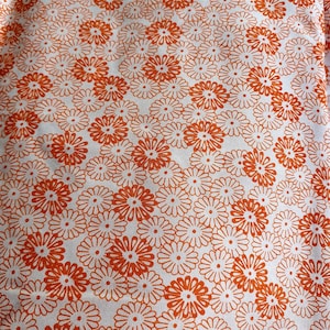 Op de afbeelding: Close-up van een witte stof met een herhaald patroon van oranje madeliefachtige bloemen. De bloemen hebben een centrale cirkel met uitstralende bloemblaadjes, waardoor een dicht, gestructureerd ontwerp ontstaat. Het algehele effect is een helder, bloemenpatroon.