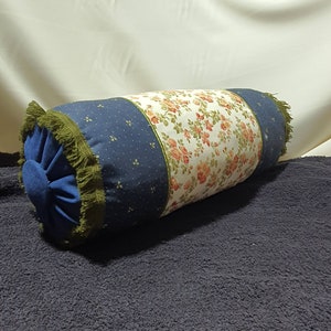 May include: A blue and white floral patterned bolster pillow with green fringe trim. The pillow is filled with a soft, comfortable material.