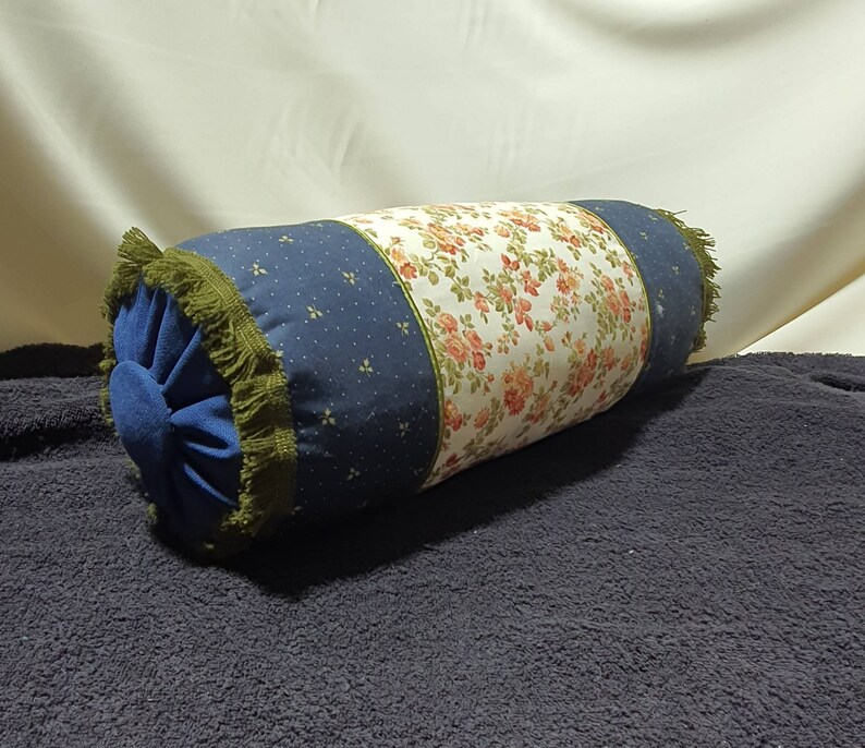 May include: A blue and white floral patterned bolster pillow with green fringe trim. The pillow is filled with a soft, comfortable material.
