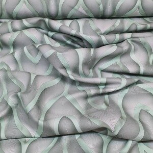 Może przedstawiać: Zielony materiał z białym i szarym geometrycznym wzorem. Materiał jest złożony i udrapowany, co pokazuje fakturę i wzór.