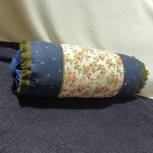 May include: A cylindrical pillow with a blue and white polka dot fabric on one side and a floral fabric on the other side. The pillow has a green fringe trim.