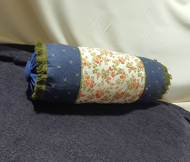 May include: A cylindrical pillow with a blue and white polka dot fabric on one side and a floral fabric on the other side. The pillow has a green fringe trim.
