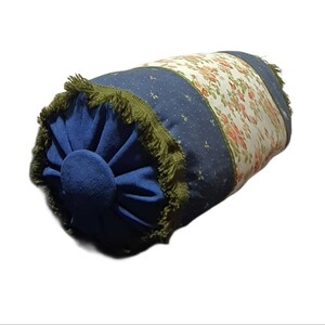 May include: A blue and white patterned cylindrical pillow with a green fringe trim. The pillow has a floral design on one side and a blue fabric with white dots on the other side.