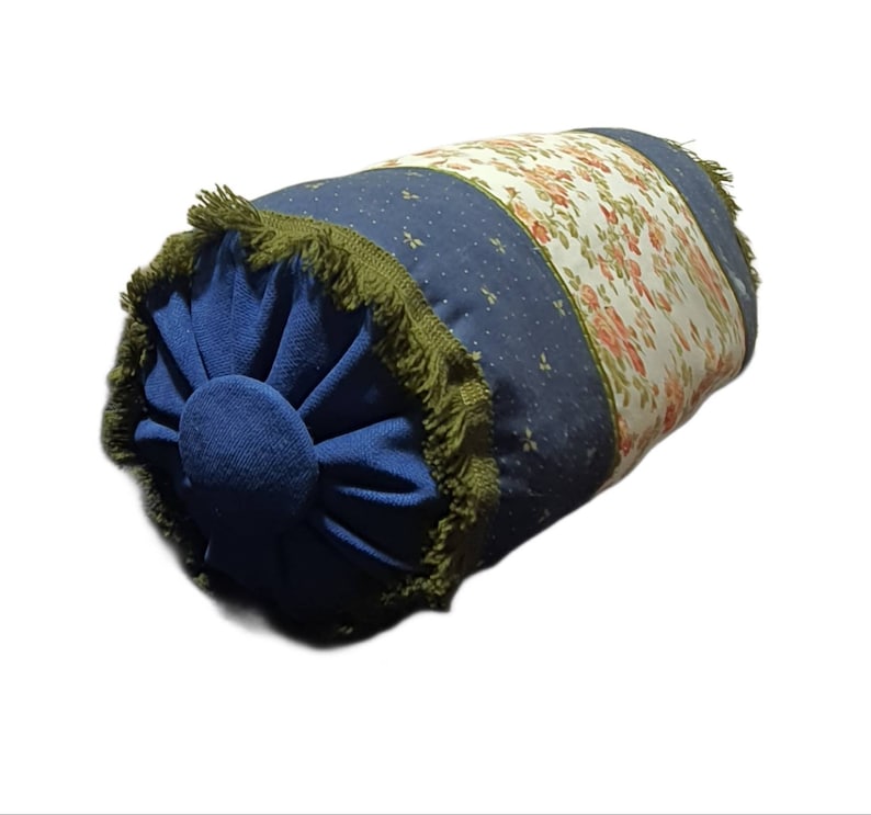 May include: A blue and white patterned cylindrical pillow with a green fringe trim. The pillow has a floral design on one side and a blue fabric with white dots on the other side.