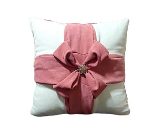 May include: A white pillow with a large pink bow and a jeweled accent. The pillow is shaped like a gift box.