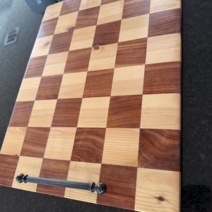 May include: A rectangular wooden serving tray with a checkerboard pattern. The squares alternate between light and dark wood tones. It has two black metal handles on the top and bottom. The tray is designed for serving food or displaying items.