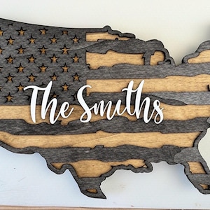 Könnte beinhalten: Wandkunst aus Holz in Form der Vereinigten Staaten, mit Flaggenmuster. Der obere Teil hat Sterne, die Mitte Streifen und der untere Teil ist aus Naturholz. Der Name "The Smiths" ist in Weiß quer durch die Mitte geschrieben.
