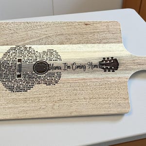 May include: Wooden cutting board with a handle, featuring a guitar design and the text "Mama, I'm Coming Home." The guitar design is filled with song lyrics. The board is light brown with a natural wood grain.